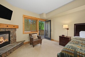 A bedroom with a stone fireplace, a wooden-framed window, a bed, a nightstand, and a chair. - White River Condo 203 at South Grand Lodge | 3BR with Partial Mt. Hood View (Government Camp)