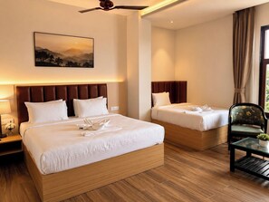 A hotel room with two beds, a ceiling fan, a nightstand with a lamp, a chair, and a window with curtains. - Daksh The Royal Braj Vrindavan (Vrindavan)