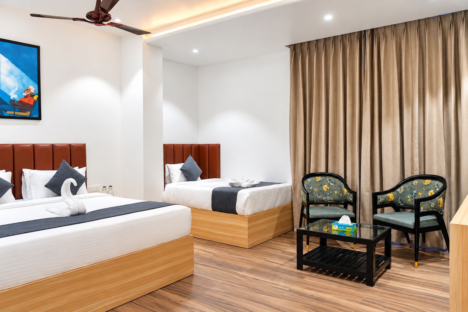 Habitación de hotel con dos camas, ventilador de techo, ventana grande con cortinas y una pequeña mesa con una silla.