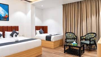 Habitación de hotel con dos camas, ventilador de techo, ventana grande con cortinas y una pequeña mesa con una silla.