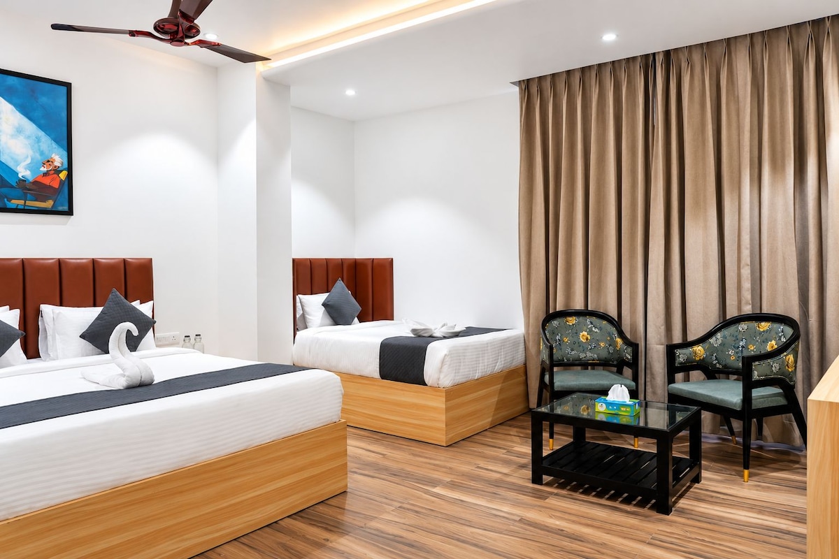 A hotel room with two beds, a ceiling fan, a painting, a window with curtains, and a small table with a chair.