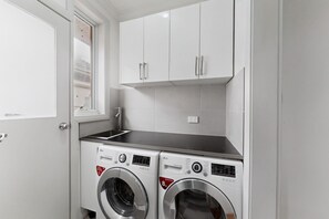 A laundry room with two front-loading washing machines, a window, and white cabinets above. - Spacious 4-Bed House with Garden in Glenroy (Glenroy)