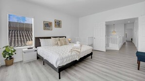 Quarto com cama, mesas de cabeceira, janela com persianas, uma planta e vista para um edifício.