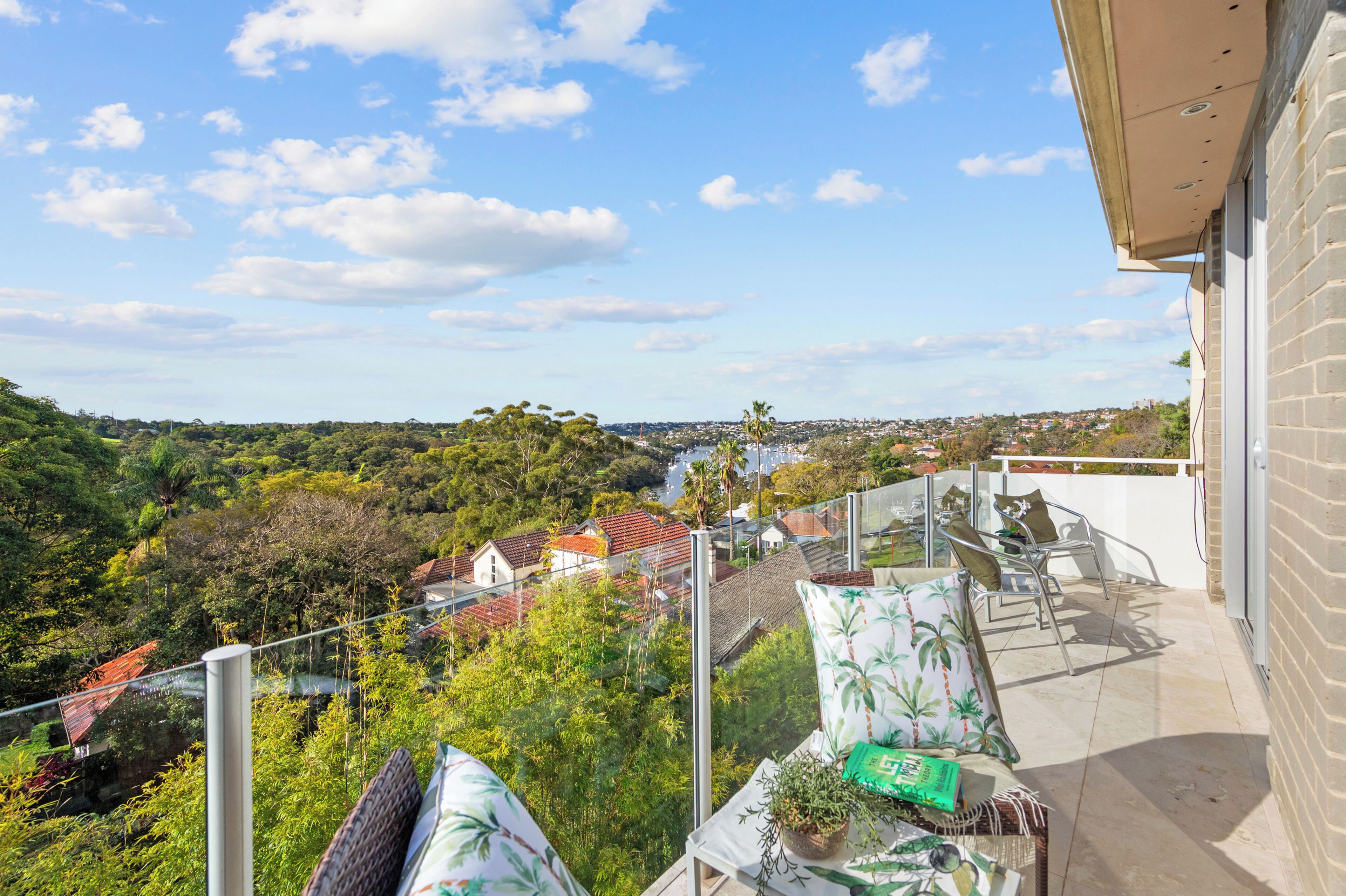 Un balcón con vistas a una zona residencial, una mesa con una planta y un libro, y una silla con cojines.