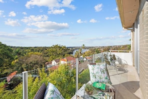 En balkon med udsigt over et boligområde, et bord med en plante og en bog, samt en stol med puder.