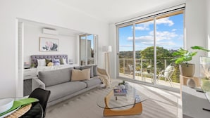 Ein modernes Wohnzimmer mit einem grauen Sofa, einem Glastisch und einem großen Fenster mit Blick ins Freie.