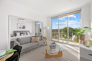 A modern living room with a grey sofa, a glass coffee table, and a large window offering a view of the outdoors. - Breathtaking Luxury 2BR | 10 Min CBD | Water Views (Cammeray)