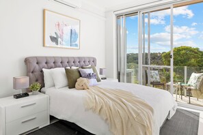 A bedroom with a large bed, a nightstand, a lamp, a framed picture, and a view of the outdoors through a sliding glass door. - Breathtaking Luxury 2BR | 10 Min CBD | Water Views (Cammeray)