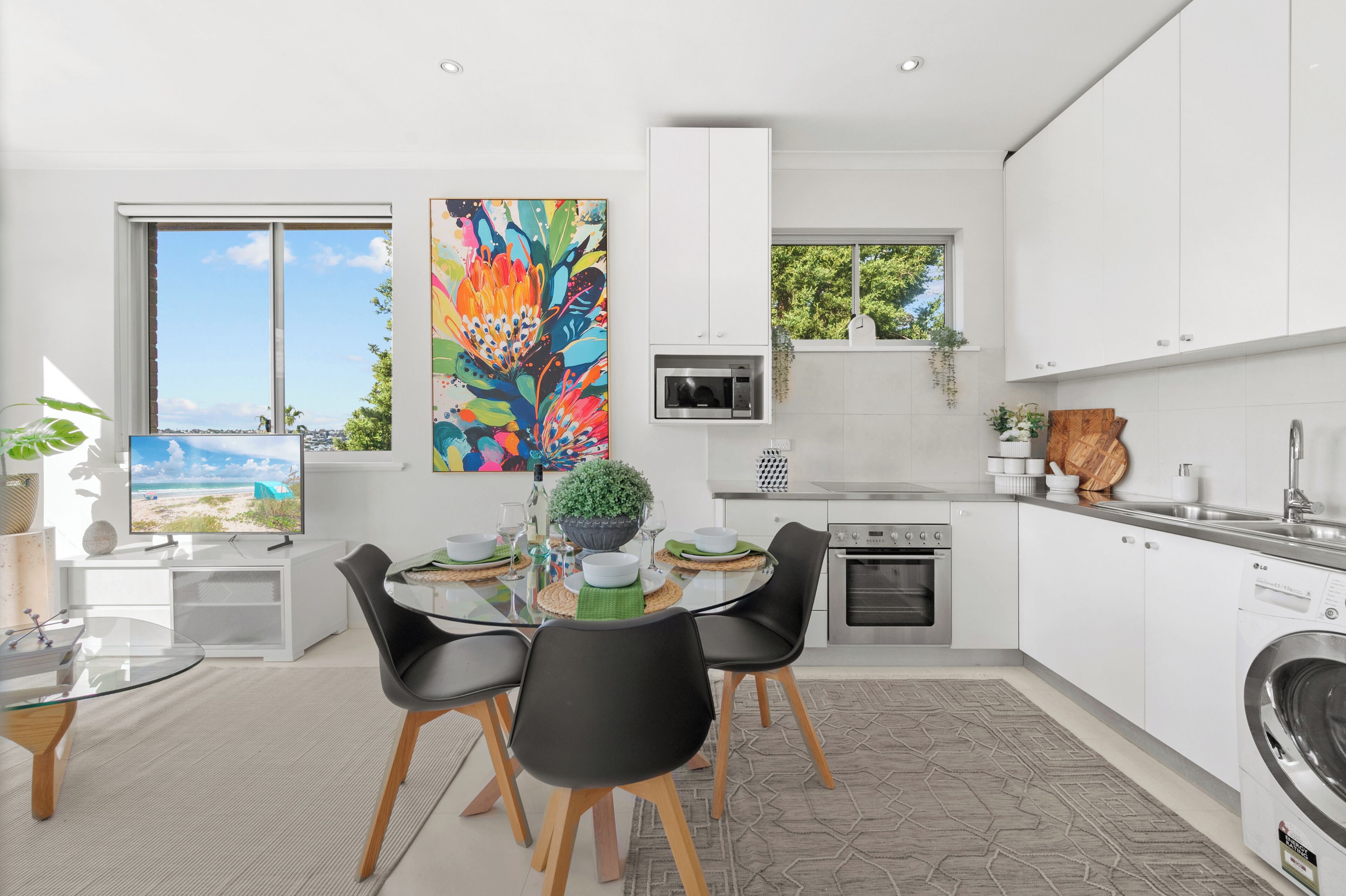 A modern kitchen with white cabinetry, a dining area with a glass table, and a large window with a beach view.