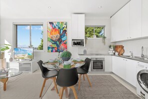 A modern kitchen with white cabinetry, a dining area with a glass table, and a large window with a beach view. - Breathtaking Luxury 2BR | 10 Min CBD | Water Views (Cammeray)