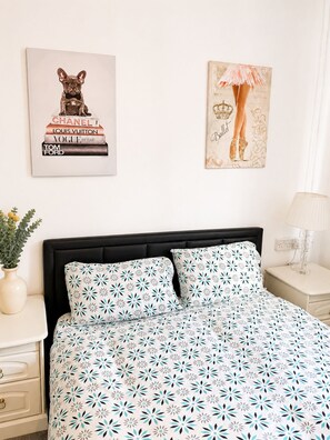 Quarto com cabeceira preta, colcha estampada com motivos florais, mesa de cabeceira com abajur e duas obras de arte nas paredes.