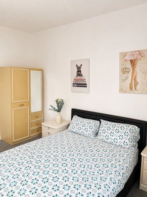 Um quarto com cama, guarda-roupa, mesa de cabeceira, um vaso com flores e duas obras de arte nas paredes.