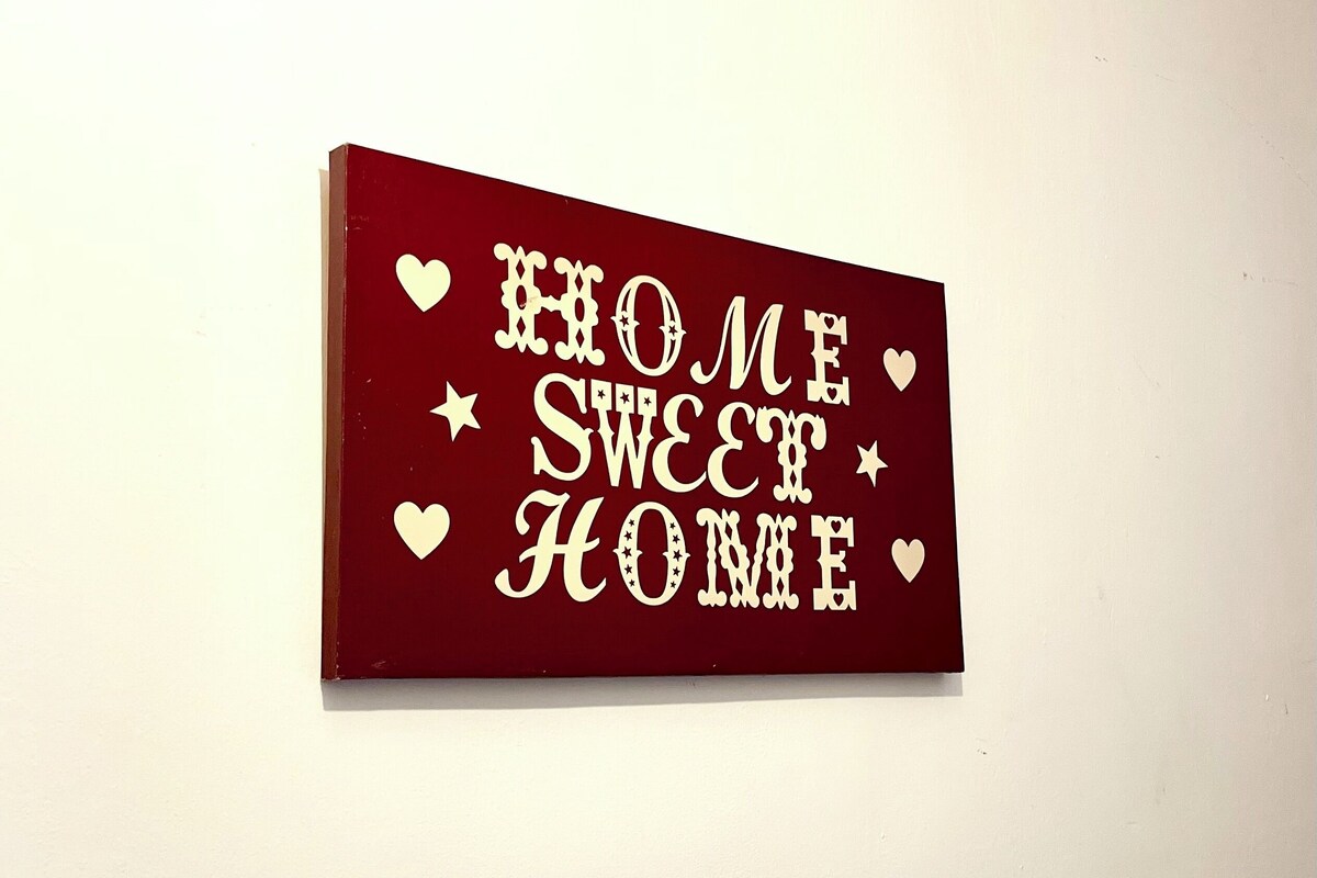 Um sinal retangular com a frase 'HOME SWEET HOME' em letras brancas sobre fundo vermelho, cercado por estrelas e corações brancos.