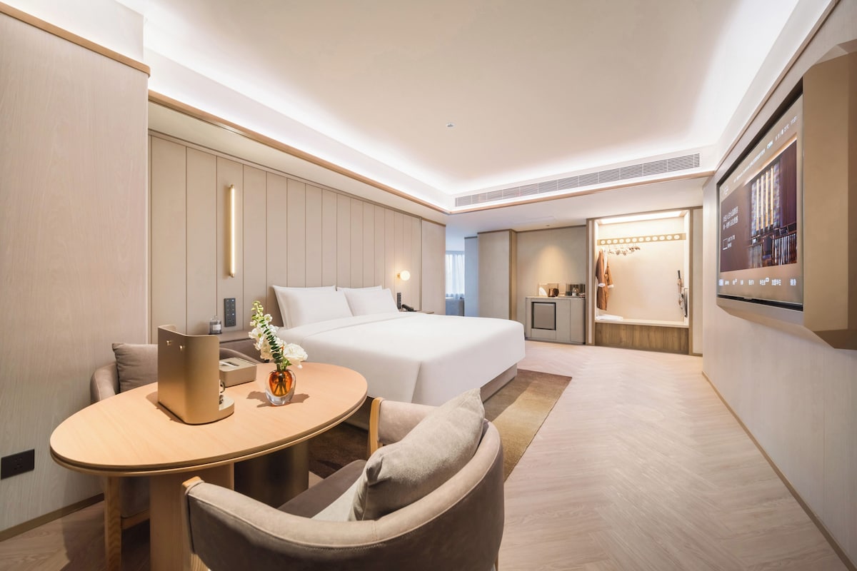A modern hotel room with a bed, bedside table, chair, and a TV screen displaying a cityscape.