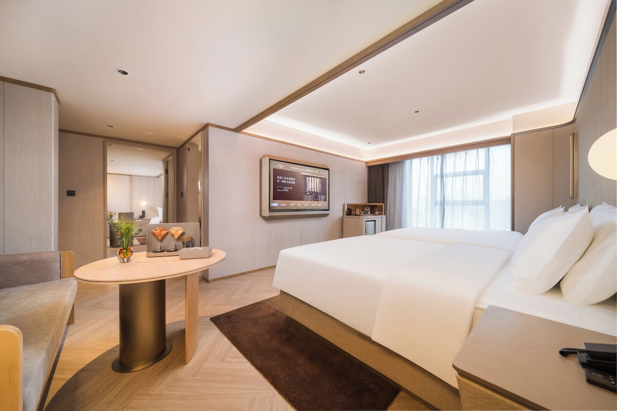 A modern hotel room with a large bed, a desk, a round table, and a television mounted on the wall.