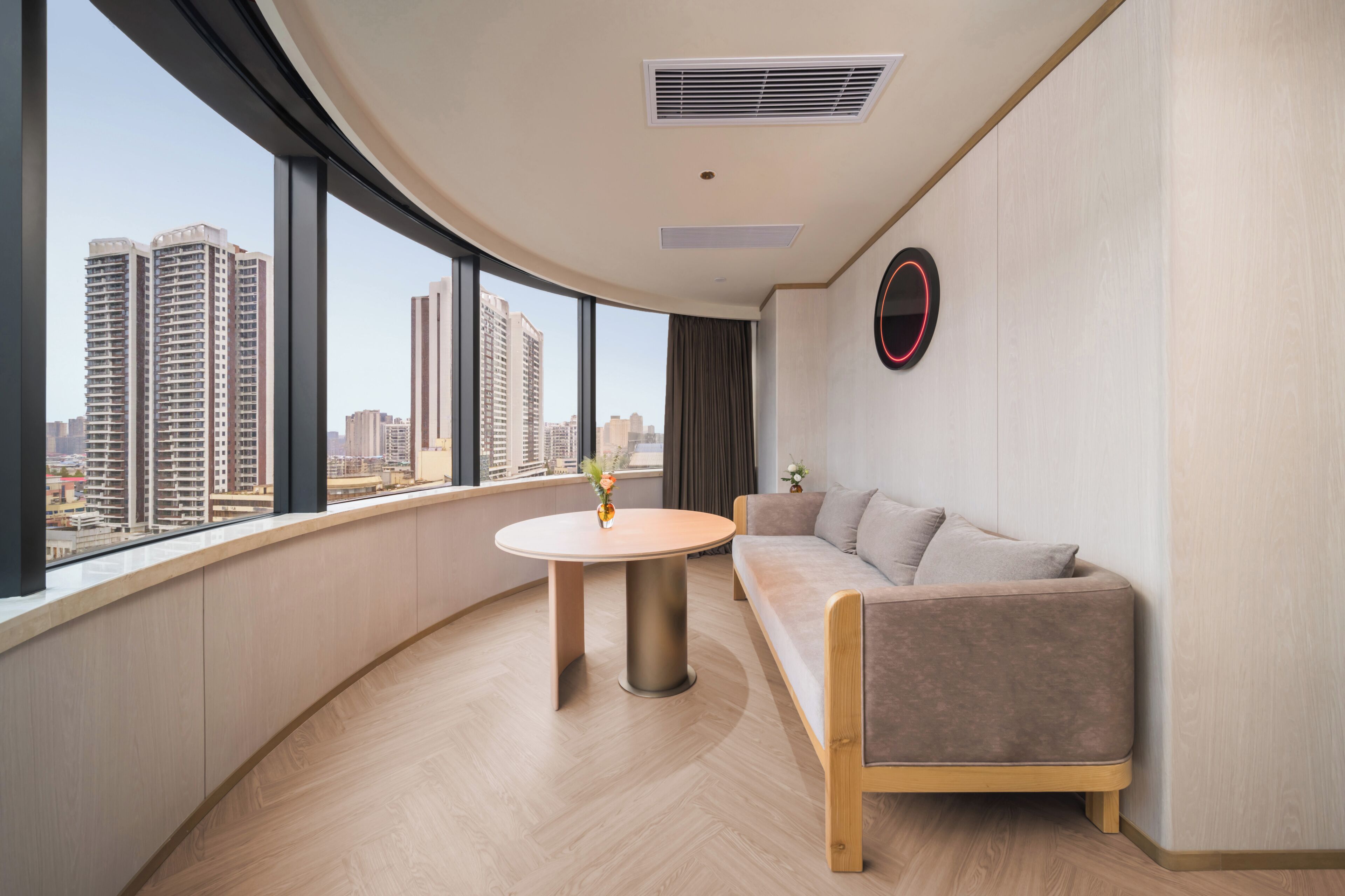 A modern hotel room with a curved balcony, a round table, a sofa, and a city view.