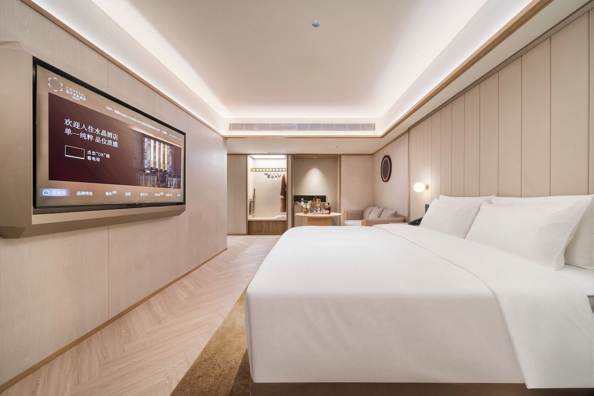 A hotel room with a large flat-screen TV mounted on the wall, a bed with white linens, a bedside table with a lamp, and a small seating area with a table and chairs.