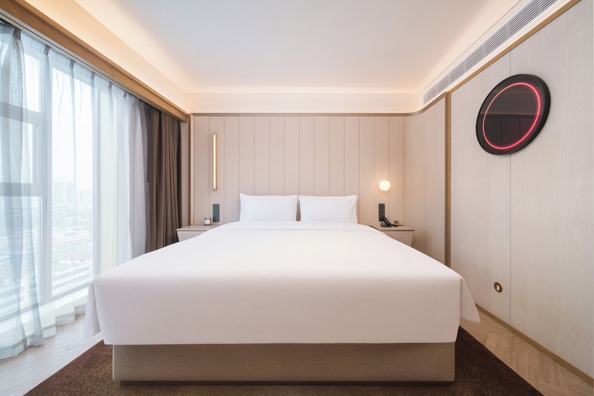 A hotel room with a large bed, a bedside table, a nightstand, a round wall clock, and a window with curtains.