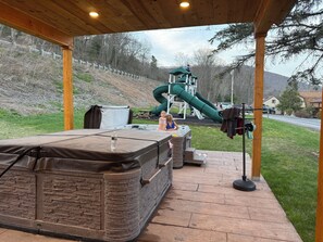 Una vasca idromassaggio con rivestimento in pietra, un pontile in legno e vista su un'area giochi con uno scivolo.