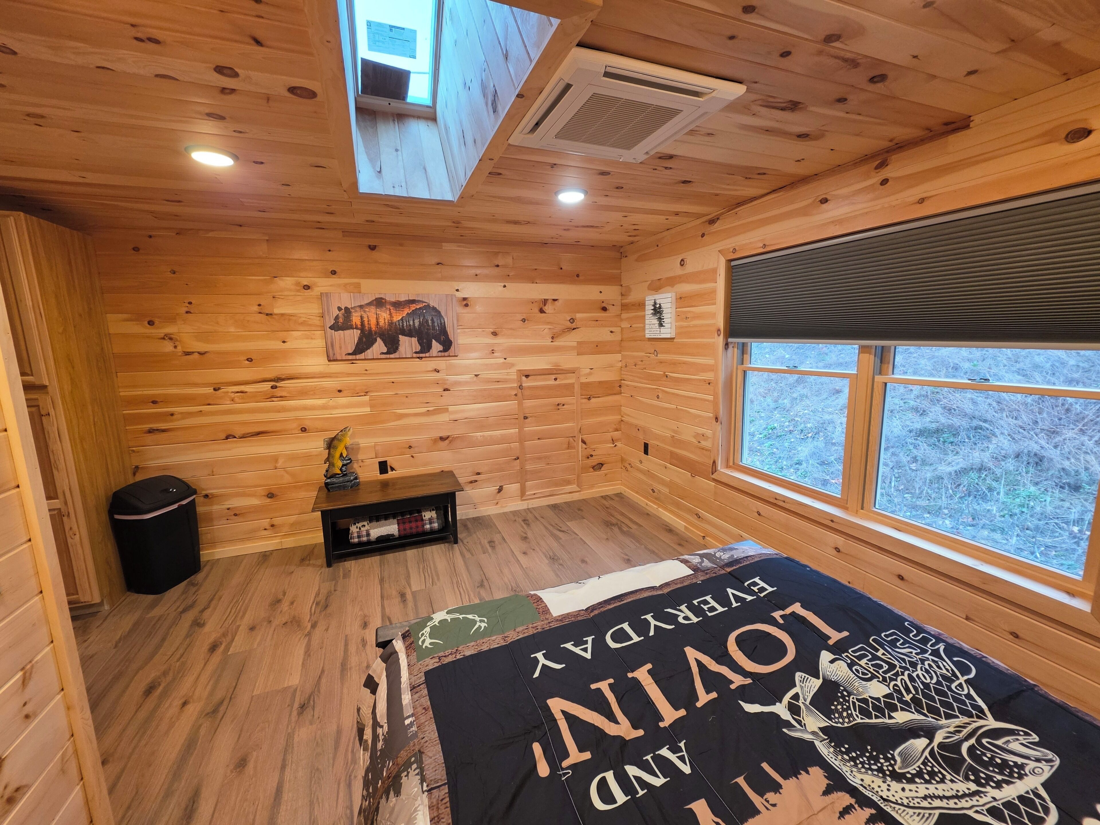 Quarto de cabana com paredes de madeira, cama com colcha temática de urso, janela com vista para árvores e um respiradouro no teto.
