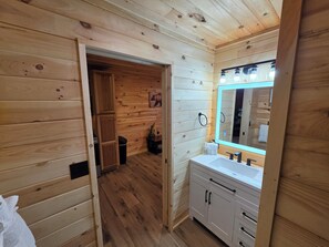 Interno di una baita in legno, con pavimento in legno, un lavandino bianco con specchio e una porta che dà su un'altra stanza.
