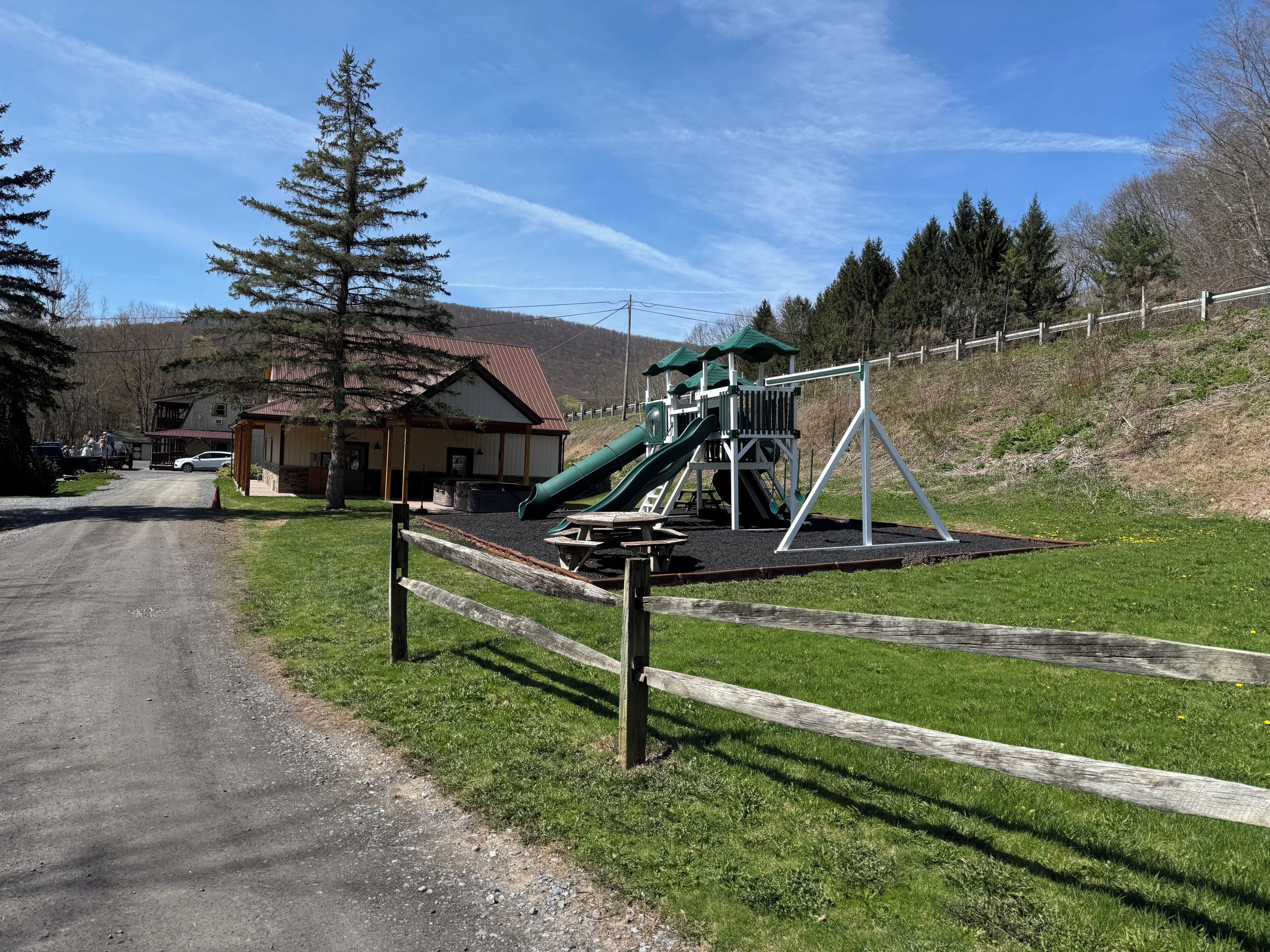 A playground with a slide and swings, a building with a red roof, and a dirt road leading to it.