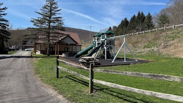 A playground with a slide and swings, a building with a red roof, and a dirt road leading to it.