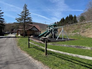 A playground with a slide and swings, a building with a red roof, and a dirt road leading to it.