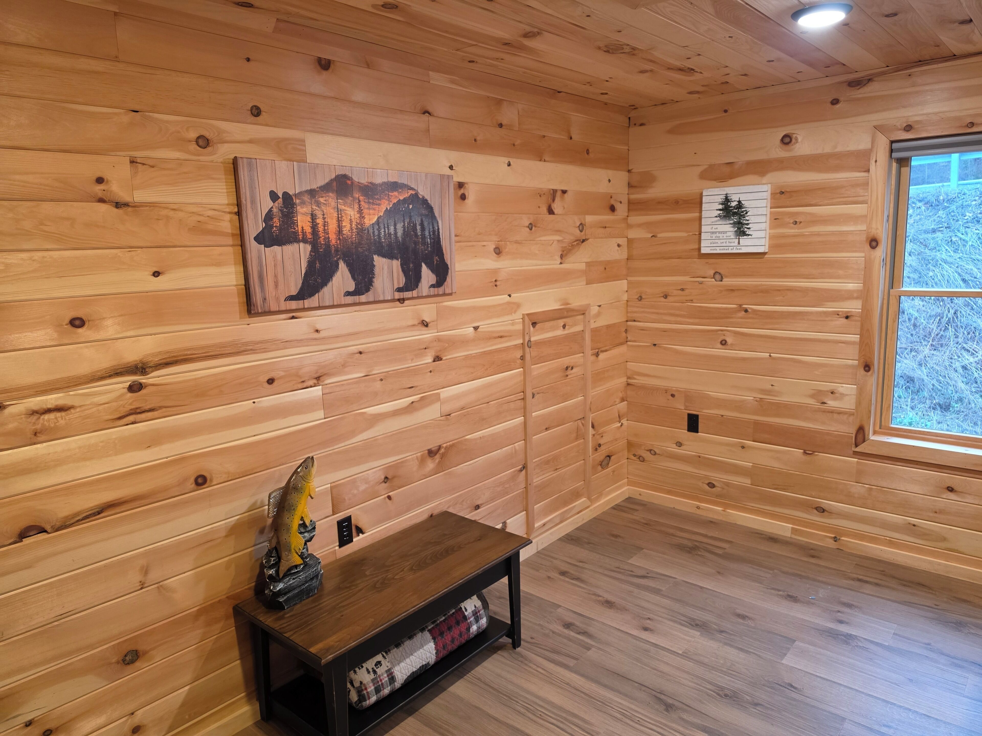 A wooden cabin interior with a bear mural, a small table with a fish sculpture, and a window with a view of trees.