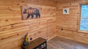 A wooden cabin interior with a bear mural, a small table with a fish sculpture, and a window with a view of trees.