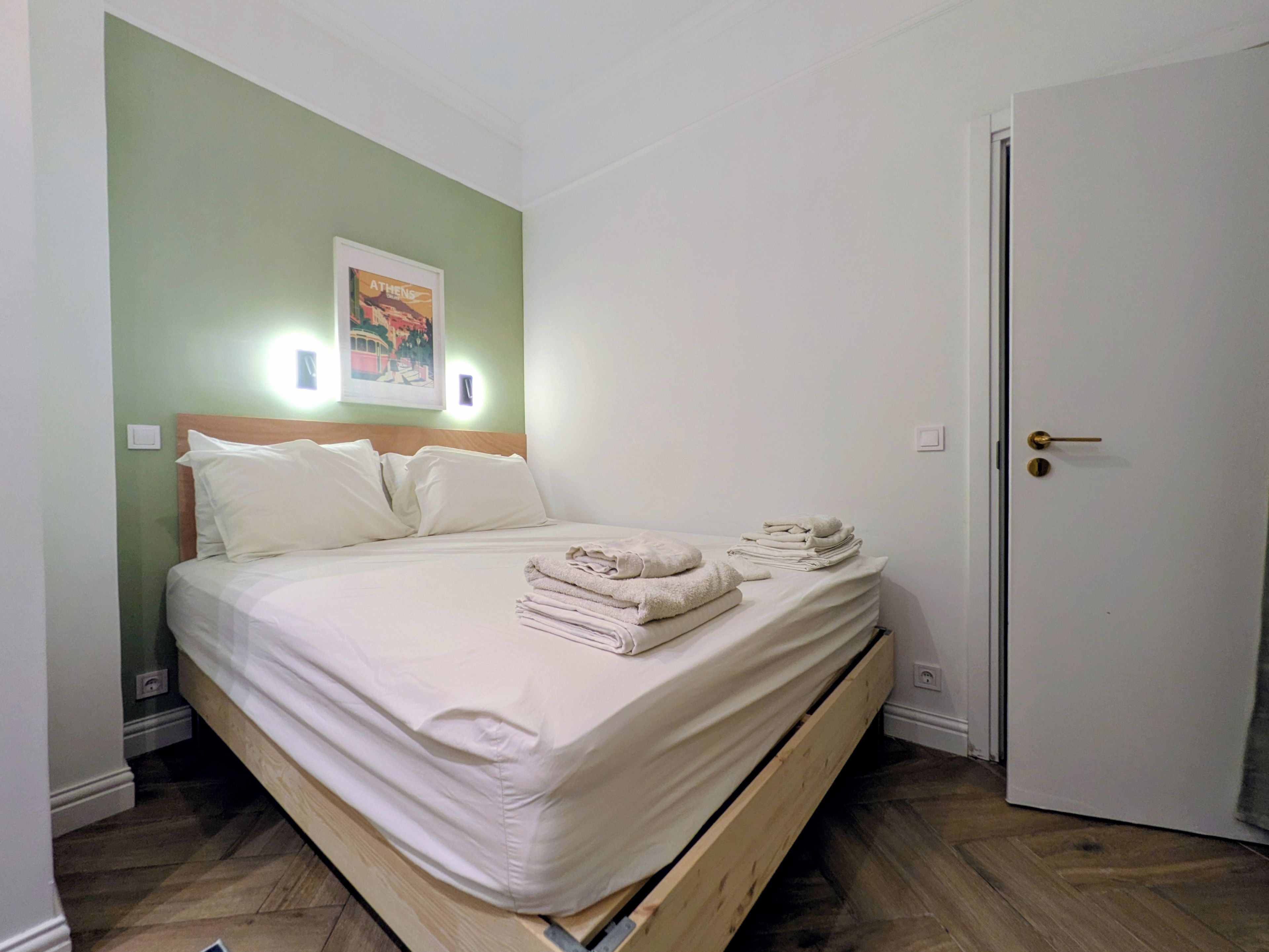 Une chambre avec un lit simple, un cadre en bois, un mur vert, un tableau encadré et une porte dotée d’une poignée en laiton.