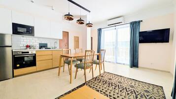 Cozinha moderna equipada com micro-ondas, forno e máquina de café. Possui área de jantar com mesa e cadeiras. Uma televisão está fixada na parede.