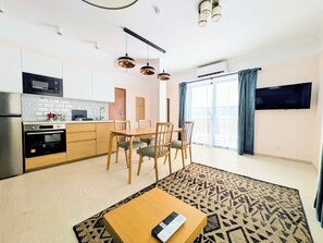 Cozinha moderna equipada com micro-ondas, forno e máquina de café. Possui área de jantar com mesa e cadeiras. Uma televisão está fixada na parede.