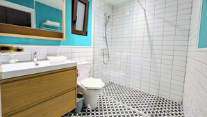 Un baño con lavamanos blanco, tocador de madera, inodoro, ducha con puerta de vidrio y suelo de baldosas con diseño geométrico.