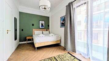 Quarto com cama, mesa de cabeceira, luminária pendente, janela com cortinas e tapete estampado.