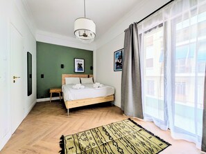 Quarto com cama, mesa de cabeceira, luminária pendente, janela com cortinas e tapete estampado.