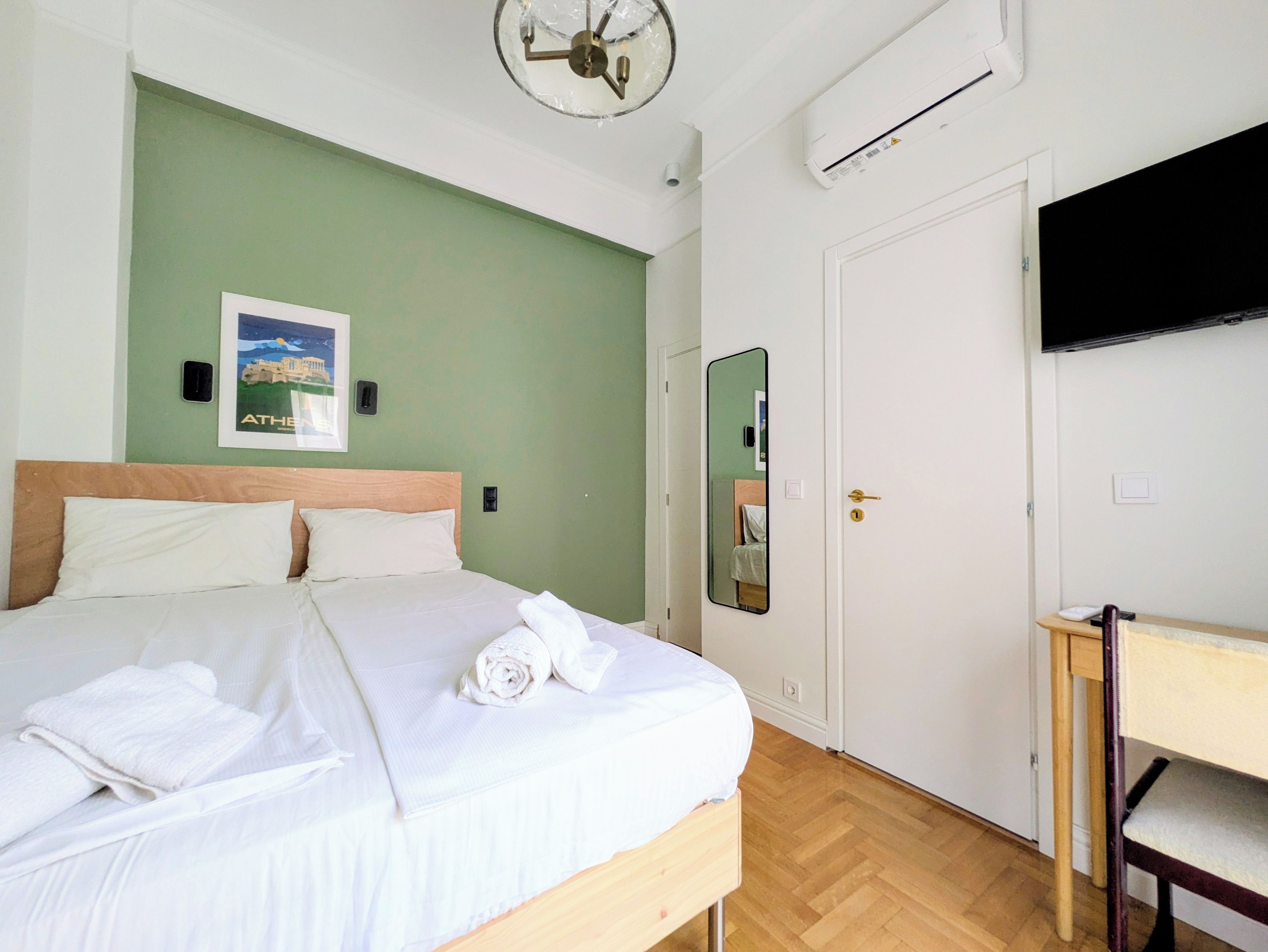 Een hotelkamer met een bed, een bureau, een stoel, een flatscreen-tv, een airconditioner, een deur en een spiegel aan de muur.