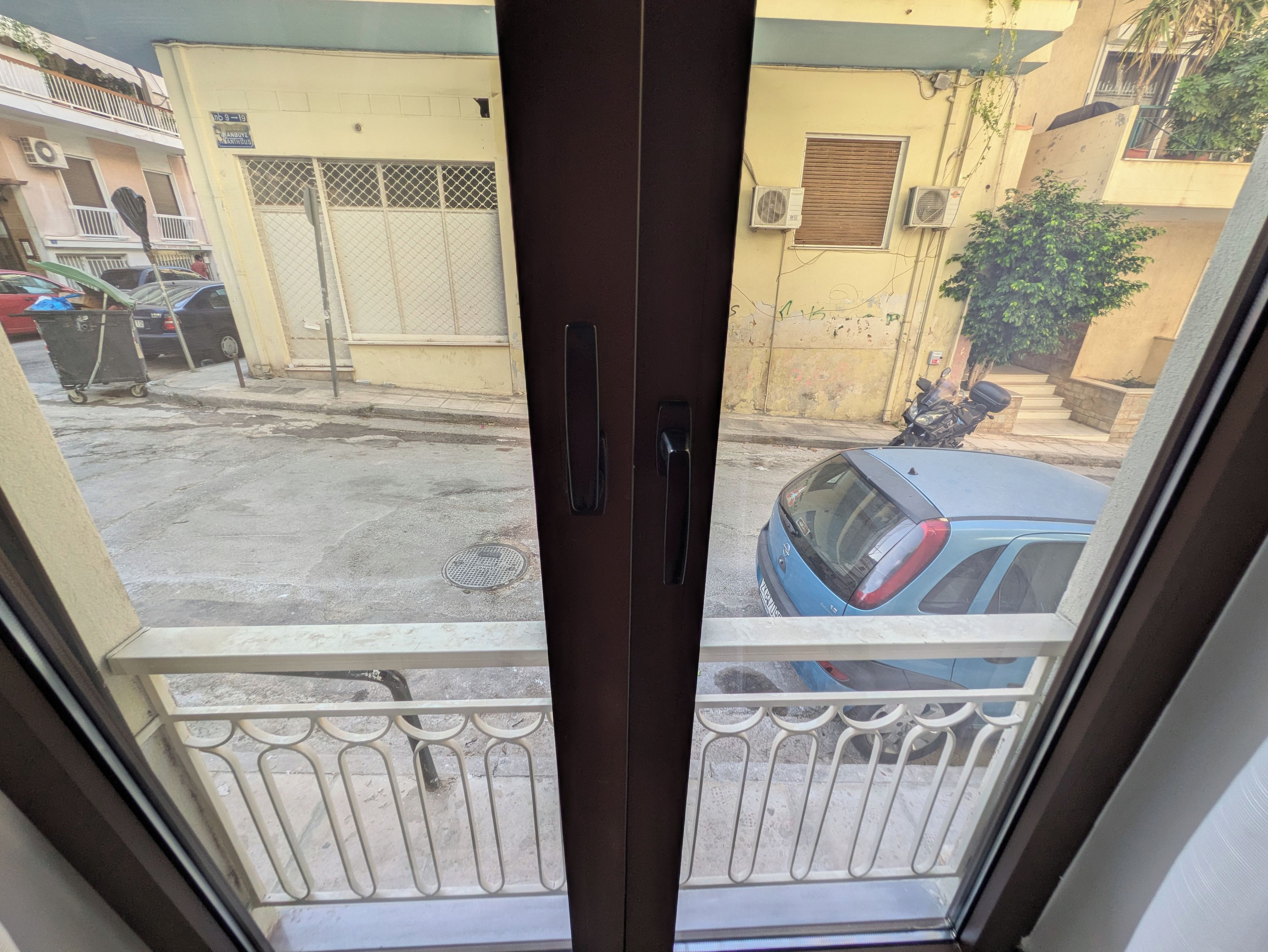 A view through a glass door with a metal railing, leading to a street with parked cars and a building with a yellow facade.