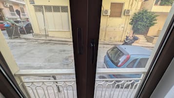 Visão através de uma porta de vidro com grade de metal, mostrando uma rua com carros estacionados e um edifício com fachada amarela.