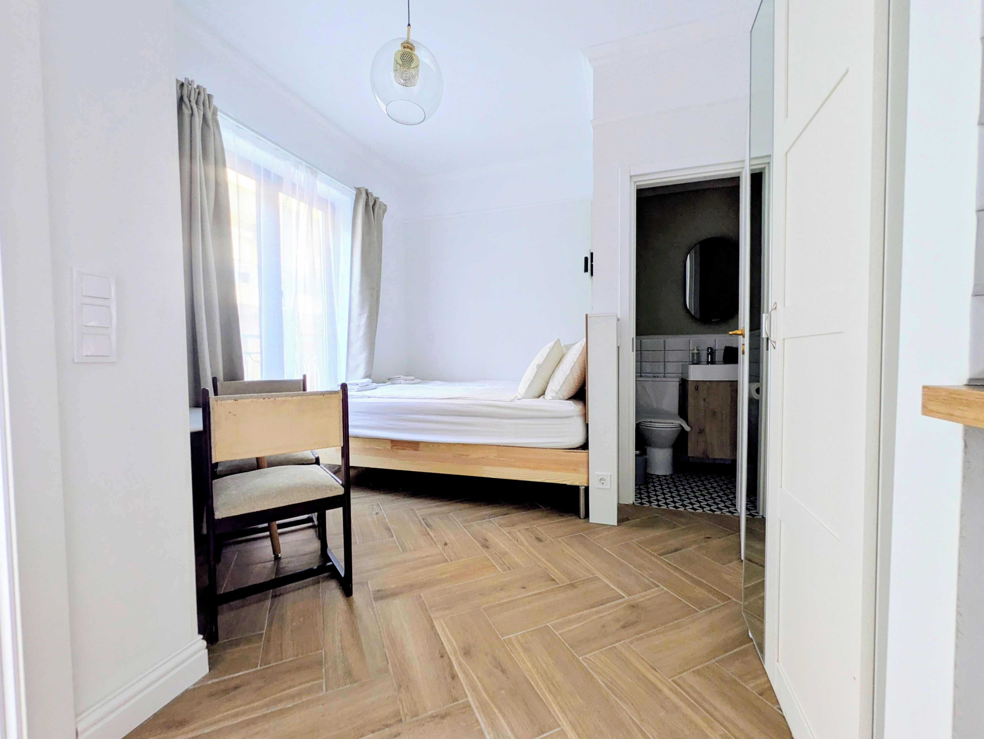 A bedroom with a bed, a chair, a window with curtains, and a bathroom visible through an open door.