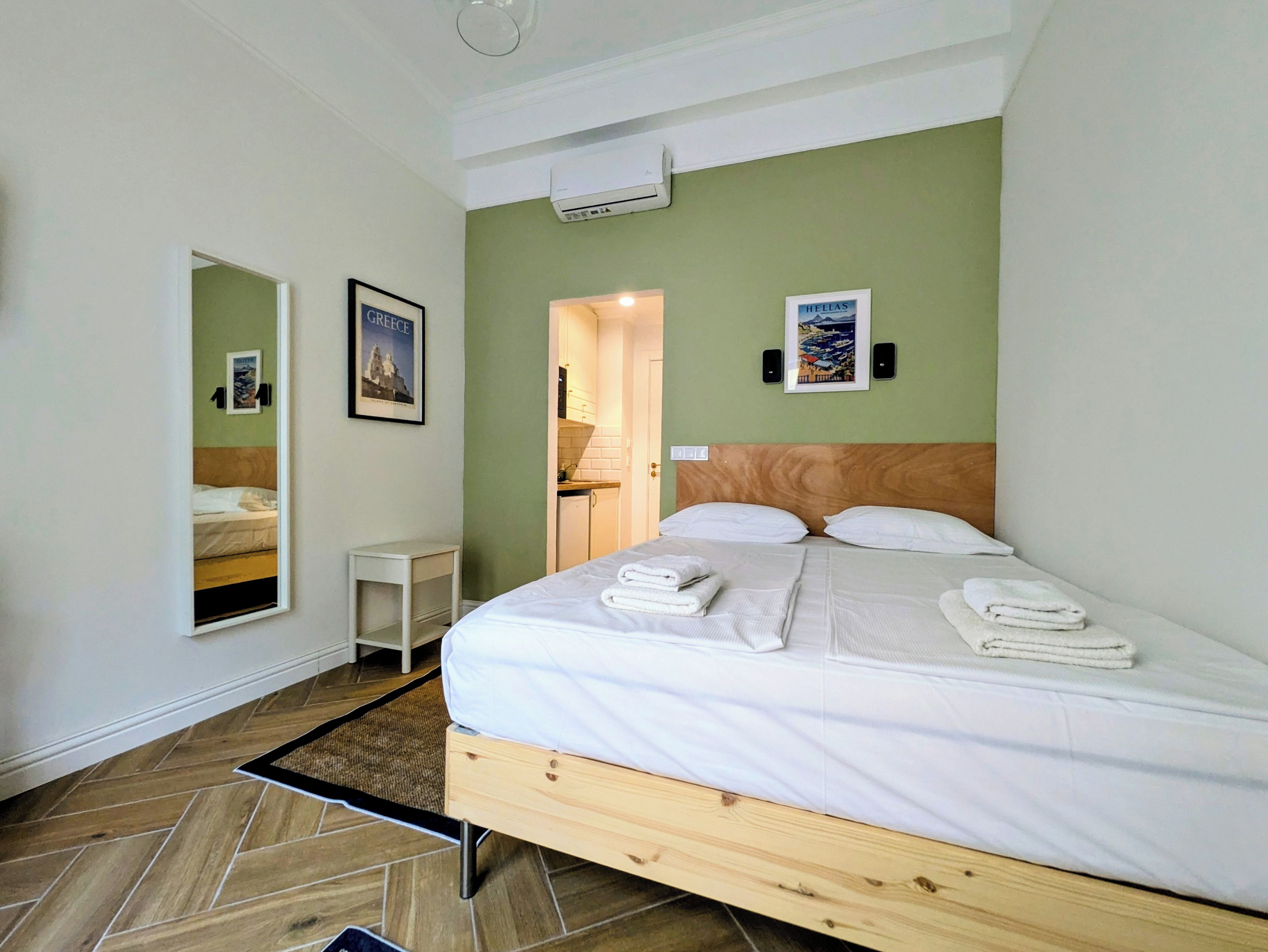 Una habitación de hotel con una cama, una mesita de noche, un espejo, un cuadro, un equipo de aire acondicionado y una puerta que da al baño.