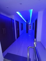 A hallway with a modern design, featuring a straight path, doors on either side, and a ceiling with blue lighting.