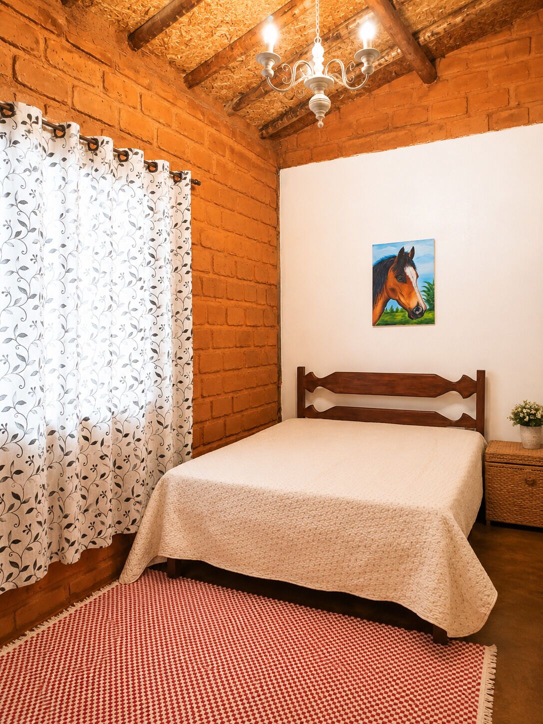 Quarto com cabeceira de madeira, cama com colcha branca texturizada, tapete quadriculado vermelho e branco, pintura de um cavalo na parede e teto de madeira com vigas aparentes.