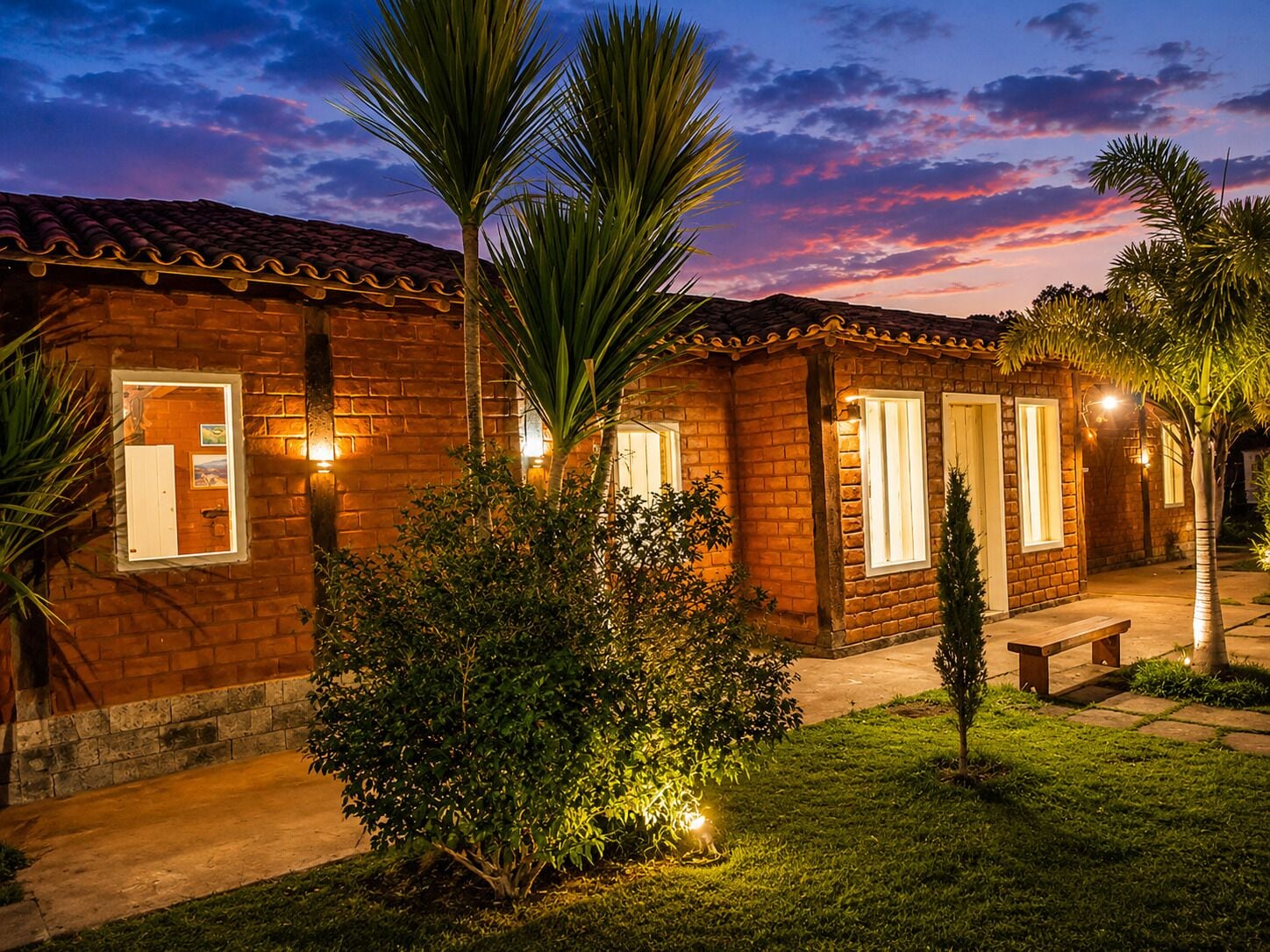 Uma casa de tijolos com telhado em cerâmica, um pequeno alpendre e uma área de jardim bem iluminada ao entardecer.