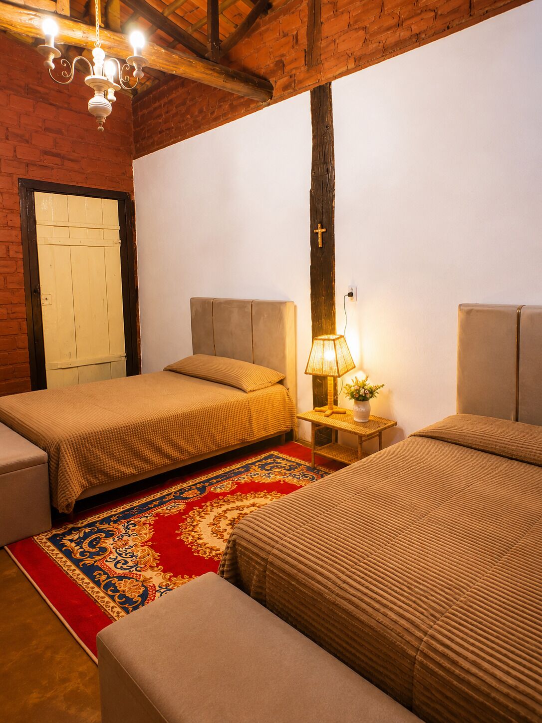 Una habitación con dos camas, un tapete rojo estampado, una cruz de madera en la pared y una lámpara en una mesita de noche.