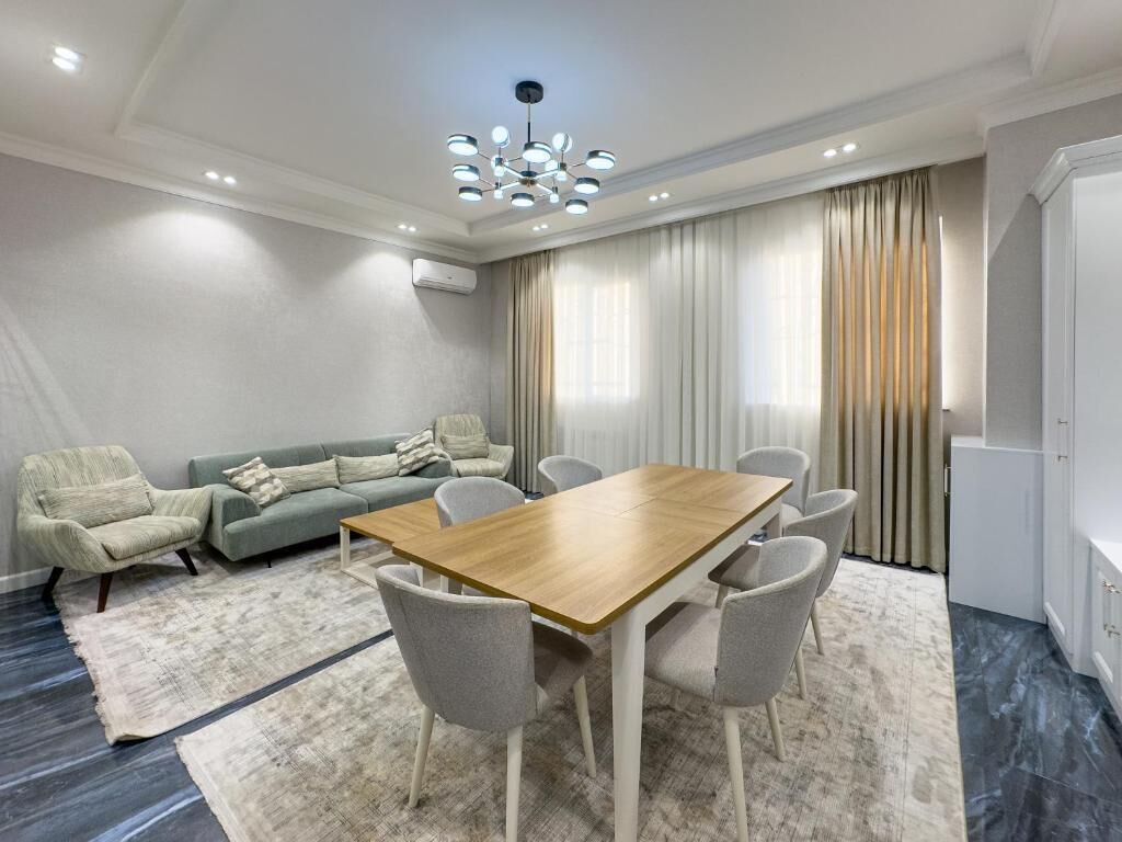 A modern living room with a wooden dining table, grey chairs, a grey sofa, and a large window with curtains.