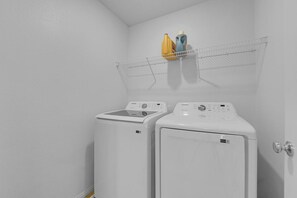 A laundry room with two white front-loading washing machines and a metal shelf above them holding detergent bottles.
