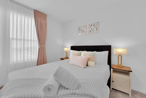 A neatly made bed with pillows, a bedside table with a lamp, and a window with curtains.