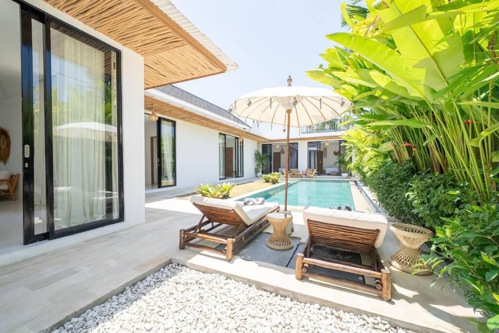 A modern outdoor pool area with lounge chairs and a large umbrella, surrounded by lush greenery and a white building with large windows.