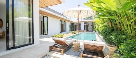A modern outdoor pool area with lounge chairs and a large umbrella, surrounded by lush greenery and a white building with large windows.
